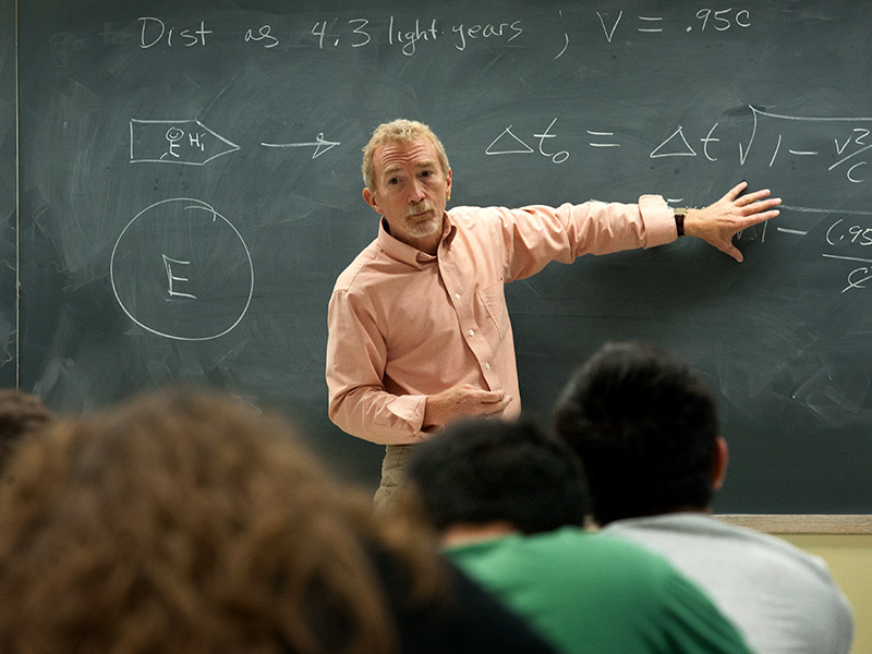 A professor stands in front of a chalk board with writing on it.