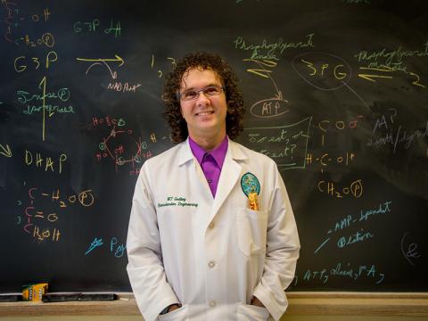 A faculty member in a lab coat stands in front of a chalkboard covered in colorful math equations. 